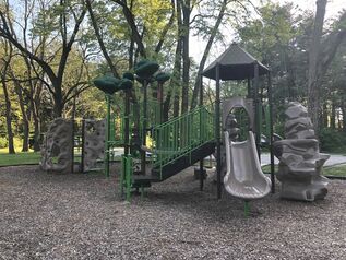 The Springfield Road Area. The playground is in a mulched area, and is complete with a climbing wall and a slide. Benches sit around the playground, and the area is surrounded with green grass and trees.